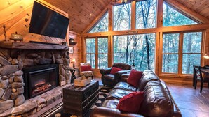A cozy cabin interior with a stone fireplace, leather sofa, and large windows offering a forest view. - Private - Creekside - Cabin + Hot Tub/Pool Table & Pet Friendly (Ellijay)