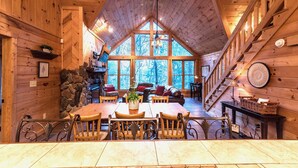 A wooden cabin interior with a dining area, stone wall, and large windows offering a view of trees. - Private - Creekside - Cabin + Hot Tub/Pool Table & Pet Friendly (Ellijay)