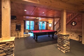 Relax in a spacious room with a pool table, wooden staircase, and large window offering a view of the surrounding trees.