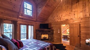 A cozy log cabin bedroom with a stone fireplace, a large bed with a quilt, and a view of the outside through a window. - Private - Creekside - Cabin + Hot Tub/Pool Table & Pet Friendly (Ellijay)