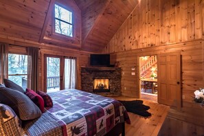 A cozy log cabin bedroom with a stone fireplace, a large bed with a quilt, and a view of the outside through a window. - Private - Creekside - Cabin + Hot Tub/Pool Table & Pet Friendly (Ellijay)