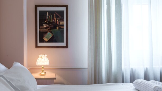 A hotel room with a neatly made bed, a bedside lamp, a framed artwork on the wall, and a window with sheer curtains. - Apartments Dobrinjska (Београд)