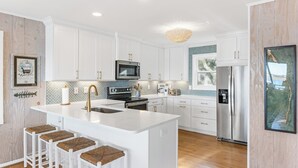 A modern kitchen with white cabinetry, stainless steel appliances, and a central island with bar stools. - The Hydeaway | Pet Friendly, Soundfront with Dock! (Topsail Beach)