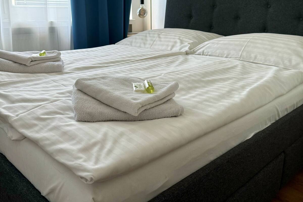 A neatly made bed with white linens and folded towels, a dark grey headboard, and a bedside lamp.