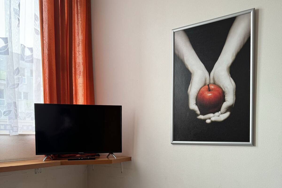 A bedroom with a TV on a wooden shelf, a bed with white bedding, and a poster of hands holding an apple on the wall.