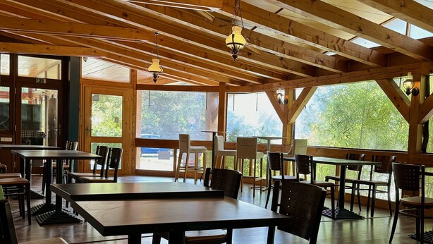 Un comedor amplio con mesas y sillas de madera, ventanas grandes que ofrecen vistas a zonas verdes y un techo alto de madera con vigas a la vista.