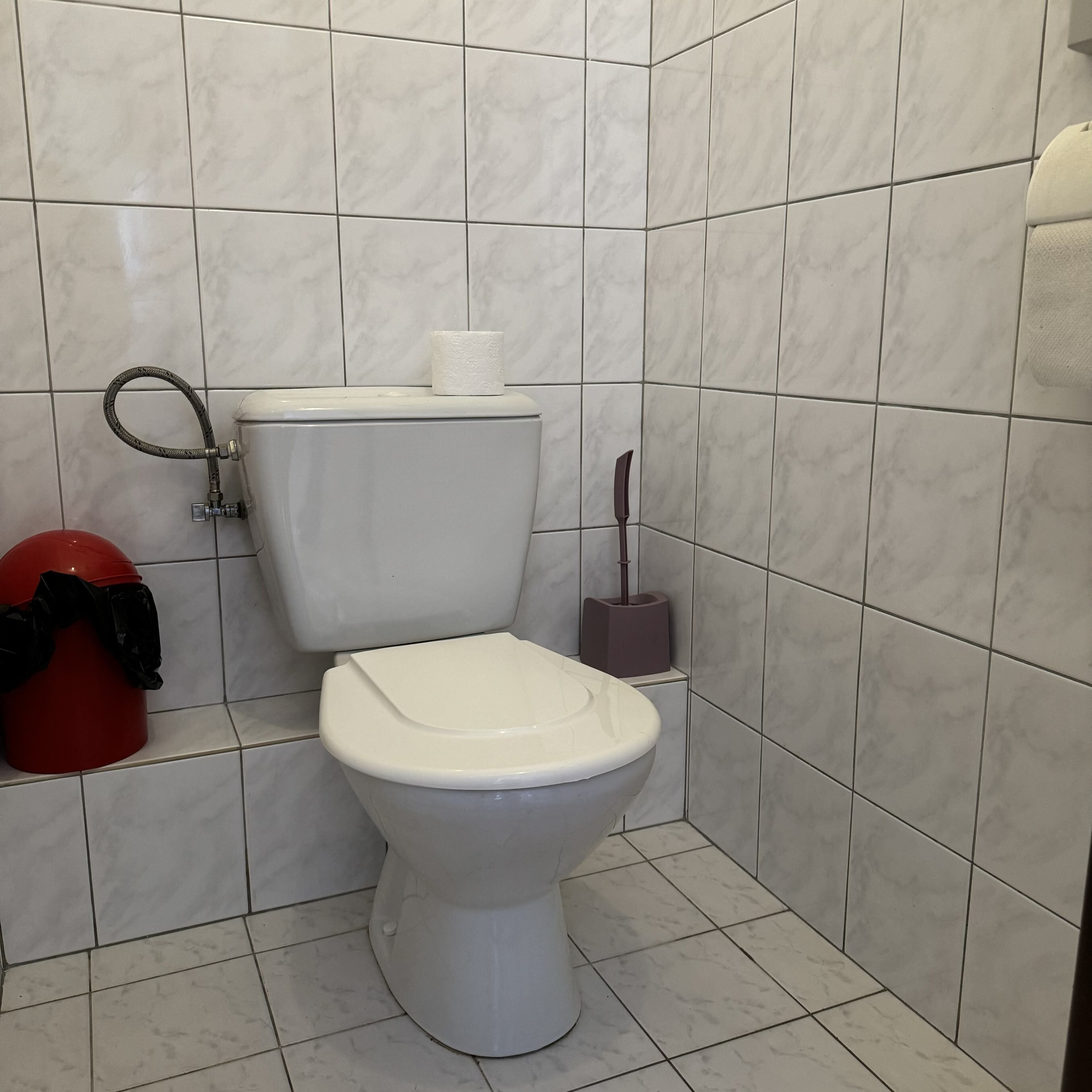 Un baño con un inodoro, un cubo rojo y un escobón.