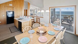 Een eetruimte met een houten tafel voor zes personen, een keuken met houten kastjes, en uitzicht op de oceaan door een schuifdeur van glas.