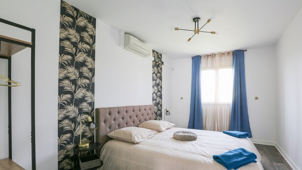 Un dormitorio con cabecera capitonada, cama con ropa de cama blanca, una toalla azul y una ventana con cortinas azules.