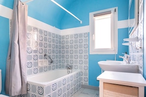 A bathroom with a blue and white tiled wall, a white bathtub, a white sink, and a window above the sink. - Le Roulis - Maison familiale avec jardin (Saint-Cast-le-Guildo)