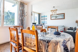 A dining area with wooden chairs, a round table covered with a newspaper print tablecloth, and a window with curtains. - Le Roulis - Maison familiale avec jardin (Saint-Cast-le-Guildo)