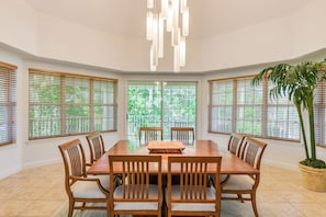 A dining room with a wooden table, six chairs, a chandelier, and large windows with blinds.