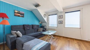 A modern living room with a blue wall, a gray sofa, a small table, and a lamp. - Callisto - Appartement Vue Mer (Luc-sur-Mer)