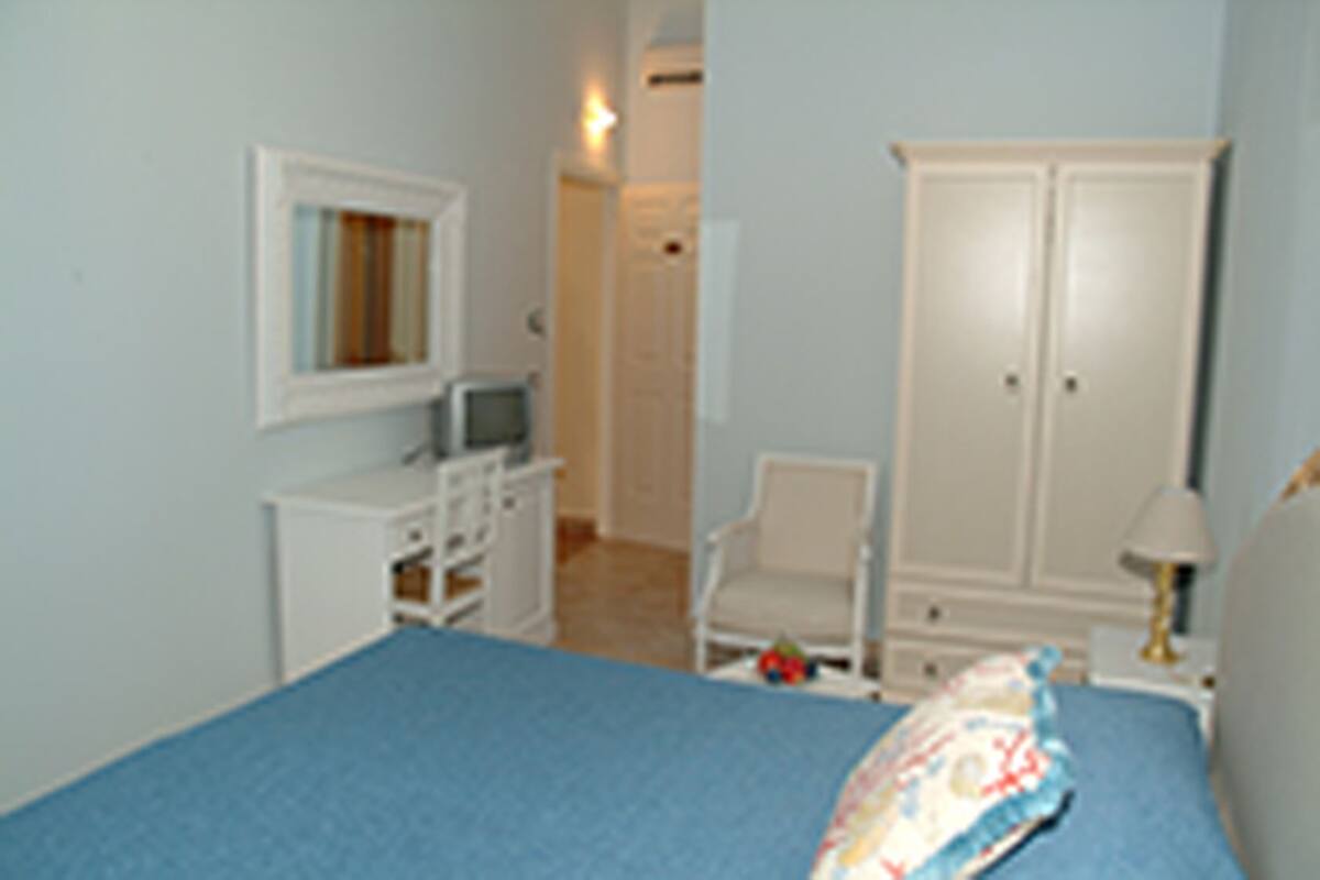 Quarto com cama, guarda-roupa, cadeira e uma mesinha de cabeceira com abajur.