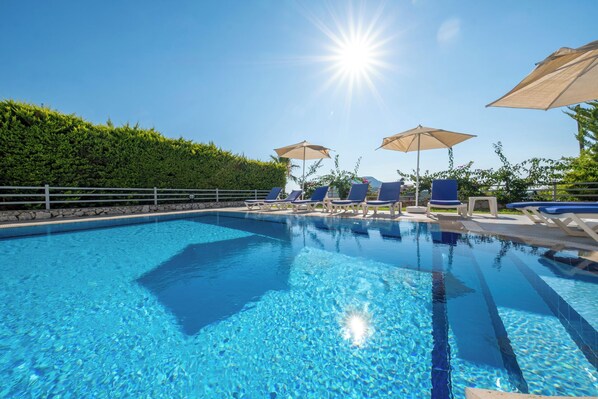 A clear blue pool with lounge chairs and umbrellas, surrounded by a hedge and a stone wall.