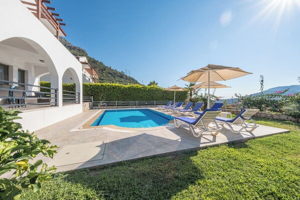 A swimming pool with lounge chairs and umbrellas, surrounded by greenery and a mountainous backdrop.