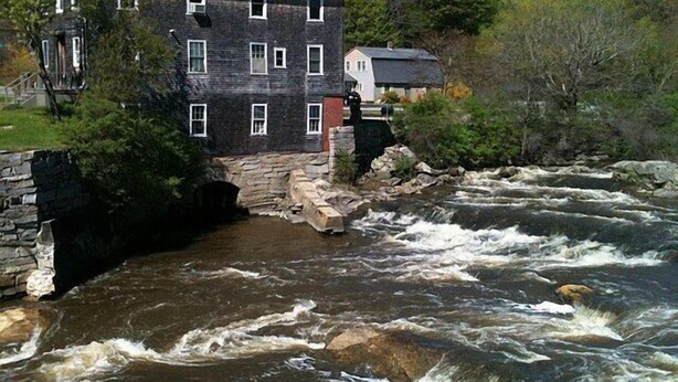 Un immeuble à plusieurs étages, doté de fondations en pierre, avec une rivière qui coule devant et une colline arborée en arrière-plan.