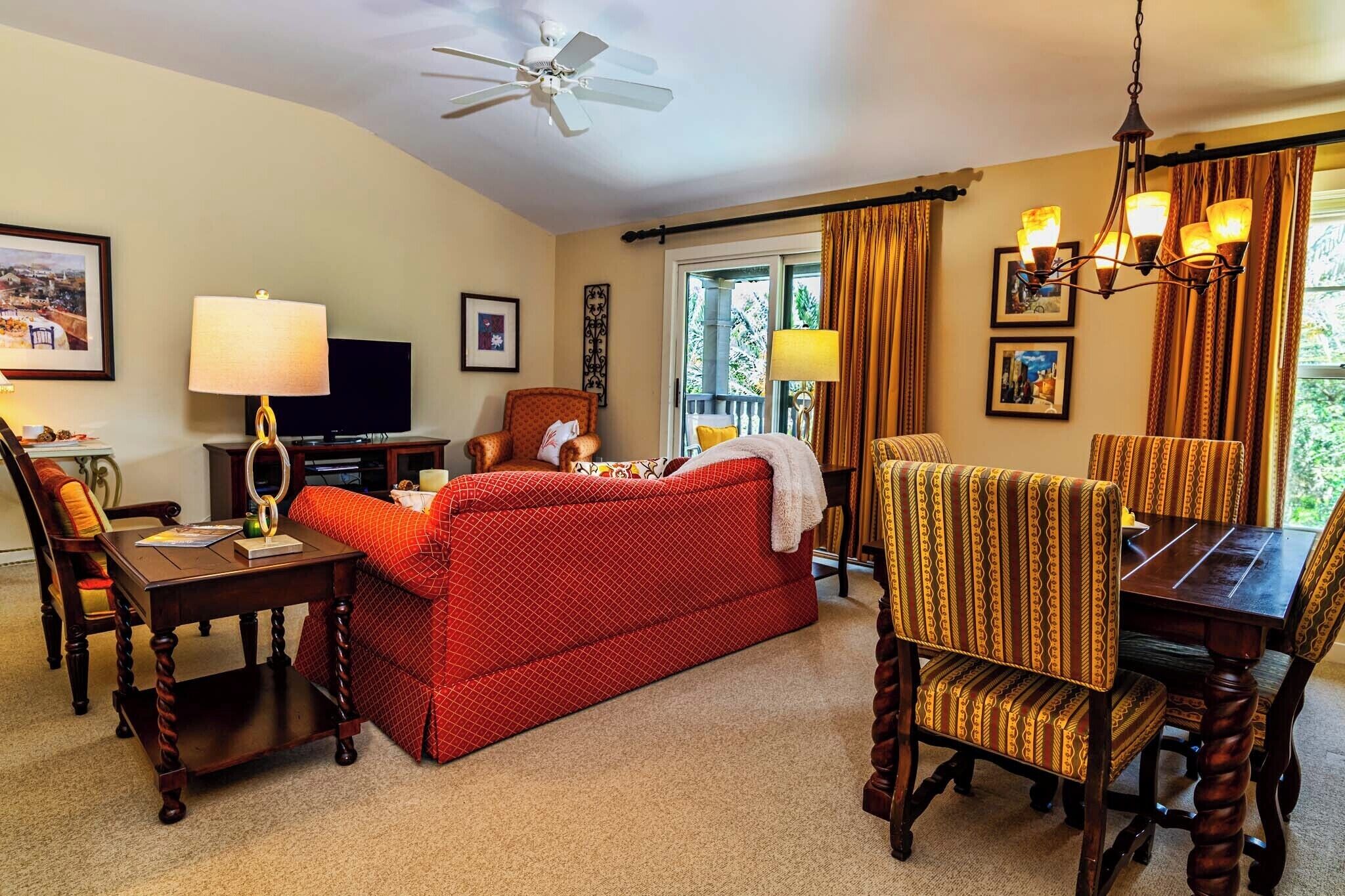 A hotel room with a red sofa, wooden dining table, and chairs, a ceiling fan, and a television.