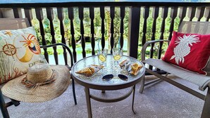 A round glass table with two chairs, a woven hat, sunglasses, and a red pillow on a balcony with a view of greenery.