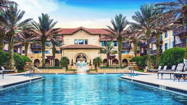 A swimming pool with a fountain, surrounded by palm trees and a building with balconies.