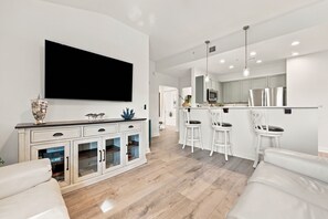 A modern living room with a flat-screen TV, a white cabinet with glass doors, and a seating area with a sofa and two white chairs.