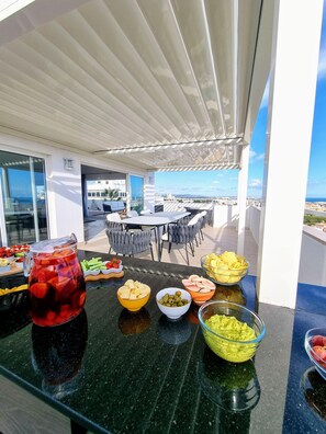 A rooftop terrace with a table set for a meal, featuring a variety of food items and a pitcher of iced tea.