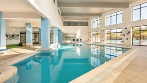 Una amplia piscina interior con agua cristalina, rodeada de paredes blancas y grandes ventanales.