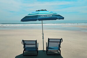 Ein Strand mit zwei leeren Stühlen und einem gestreiften Sonnenschirm mit dem Schriftzug 'Life is Good'.