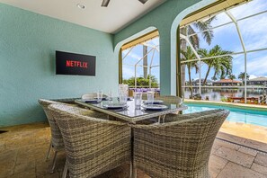 A dining area with wicker chairs, a table set for four, and a large window overlooking a pool and palm trees. - Villa Lago Vista - Waterfront Gem w/ Gulf Access (CAPE CORAL)