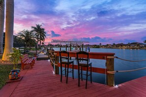 Una terrassa al costat del mar amb cadires i un banc de fusta, amb vista a una superfície d'aigua tranquil·la i una filera de cases.