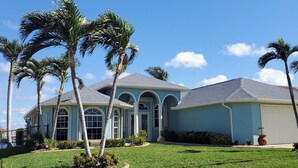 A two-story house with a light blue exterior, a grey roof, and a white garage door. There are palm trees in the front yard.