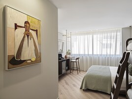 Ένα υπνοδωμάτιο με ένα κρεβάτι, ένα γραφείο, μια καρέκλα, ένα παράθυρο με λεπτά, διάφανα κουρτίνια και έναν πίνακα στον τοίχο.