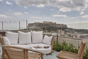 Terraço no telhado com um sofá branco, cadeiras de vime e uma mesa pequena. Ruínas antigas são visíveis ao fundo.