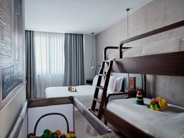 Quarto de hotel com beliches, um cercadinho infantil com brinquedos e uma janela grande com cortinas.