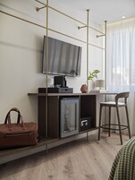Egy modern szállodai szoba, melyben a falra van szerelve egy síkképernyős televízió, található egy minibar, egy kis hűtőszekrény, egy fa íróasztal, egy szék, és a padlón egy bőr táska.
