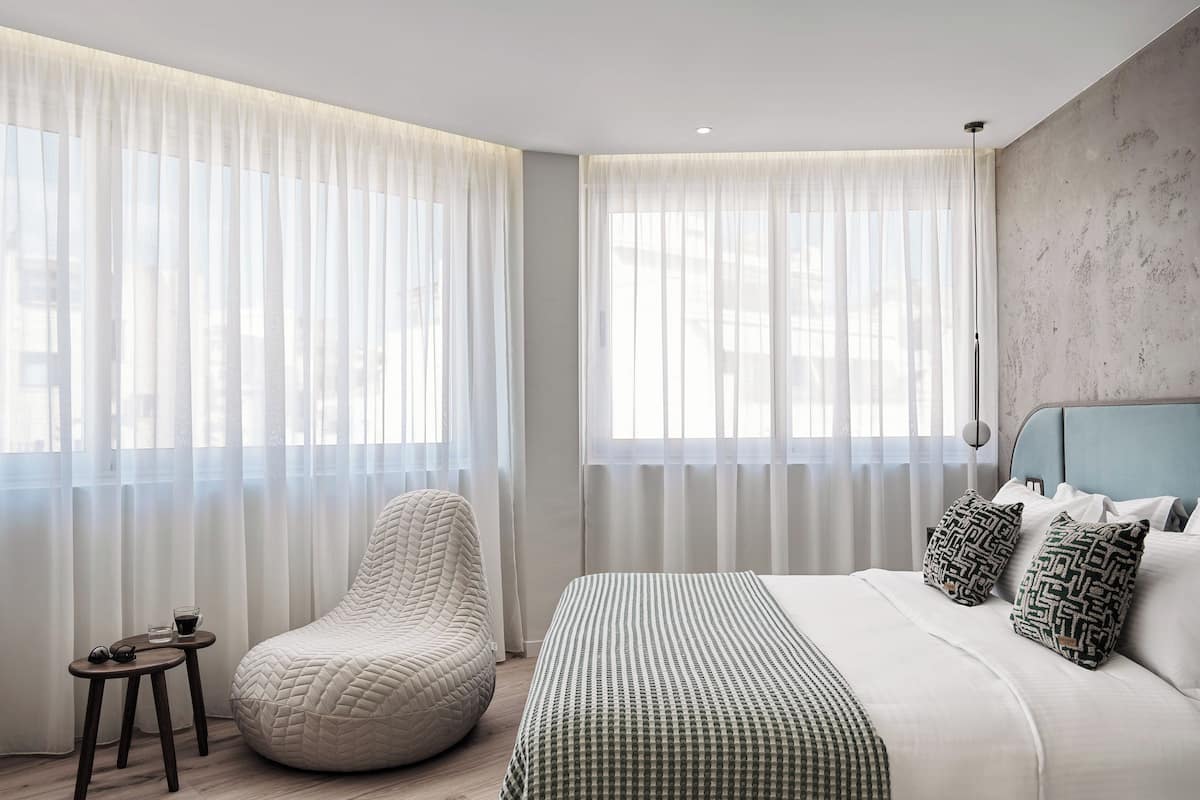 A modern bedroom with a large bed, a bean bag chair, and sheer curtains.