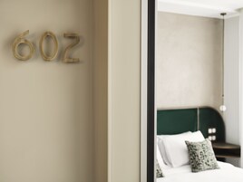 Quarto de hotel com design minimalista, com cama e cabeceira verde, roupa de cama branca, um travesseiro verde e uma placa de parede com o número '602'.
