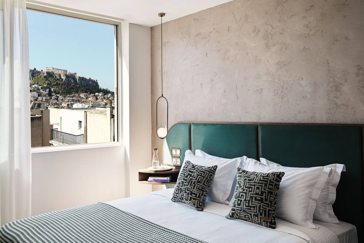 A hotel room with a large window, a green upholstered headboard, and a view of a cityscape.