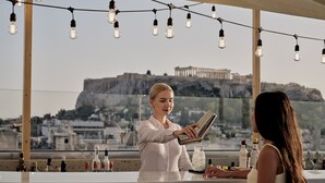 Un bar offrant une vue sur un bâtiment historique, des bouteilles d’alcool et un barman préparant un cocktail.