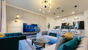 A modern living room with a sofa, a coffee table, and a television. There is a kitchen area in the background with cabinets and a refrigerator.