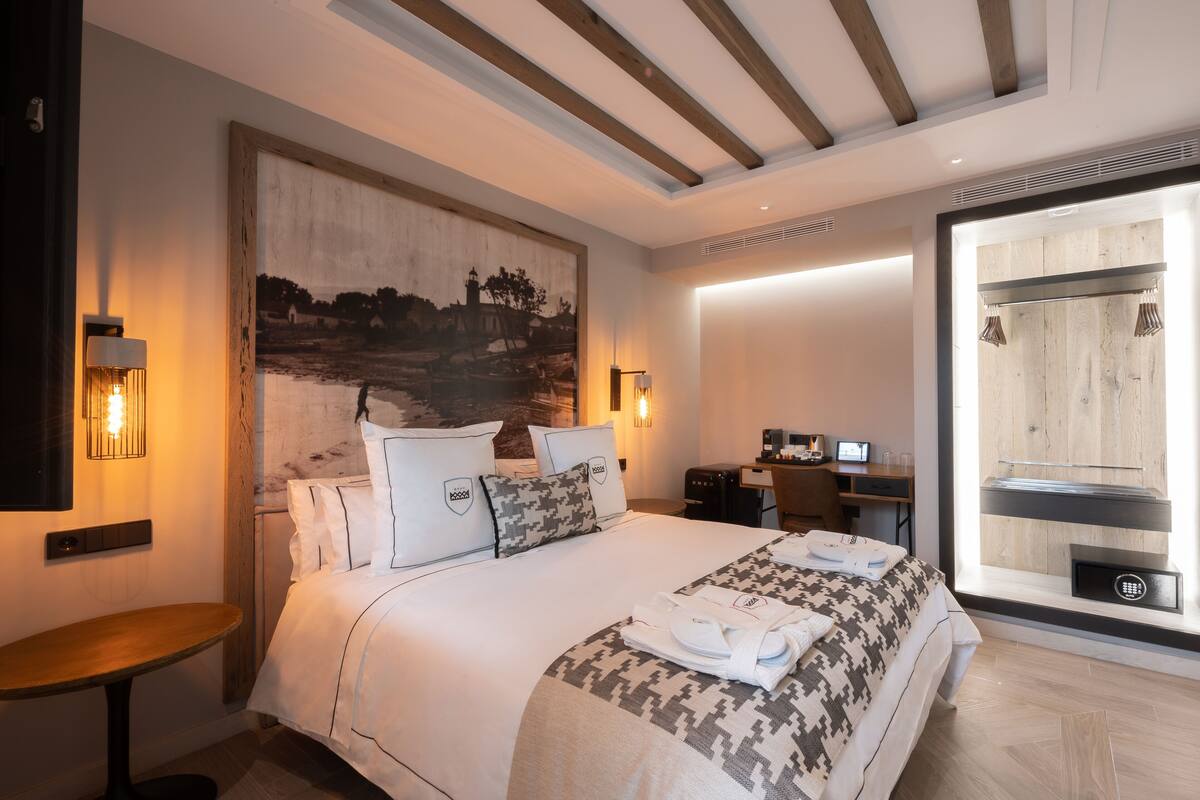 A modern hotel room with a large bed, a desk, a chair, and a large window.