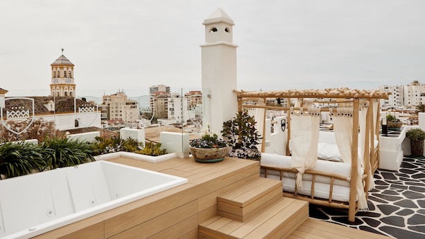 Eine Dachterrasse mit Whirlpool, einer Holzw pergola mit weißen Vorhängen und Blick auf die Stadtlandschaft, einschließlich eines hohen Turms.