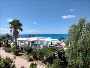 A pool area with umbrellas, palm trees, and a view of the sea.