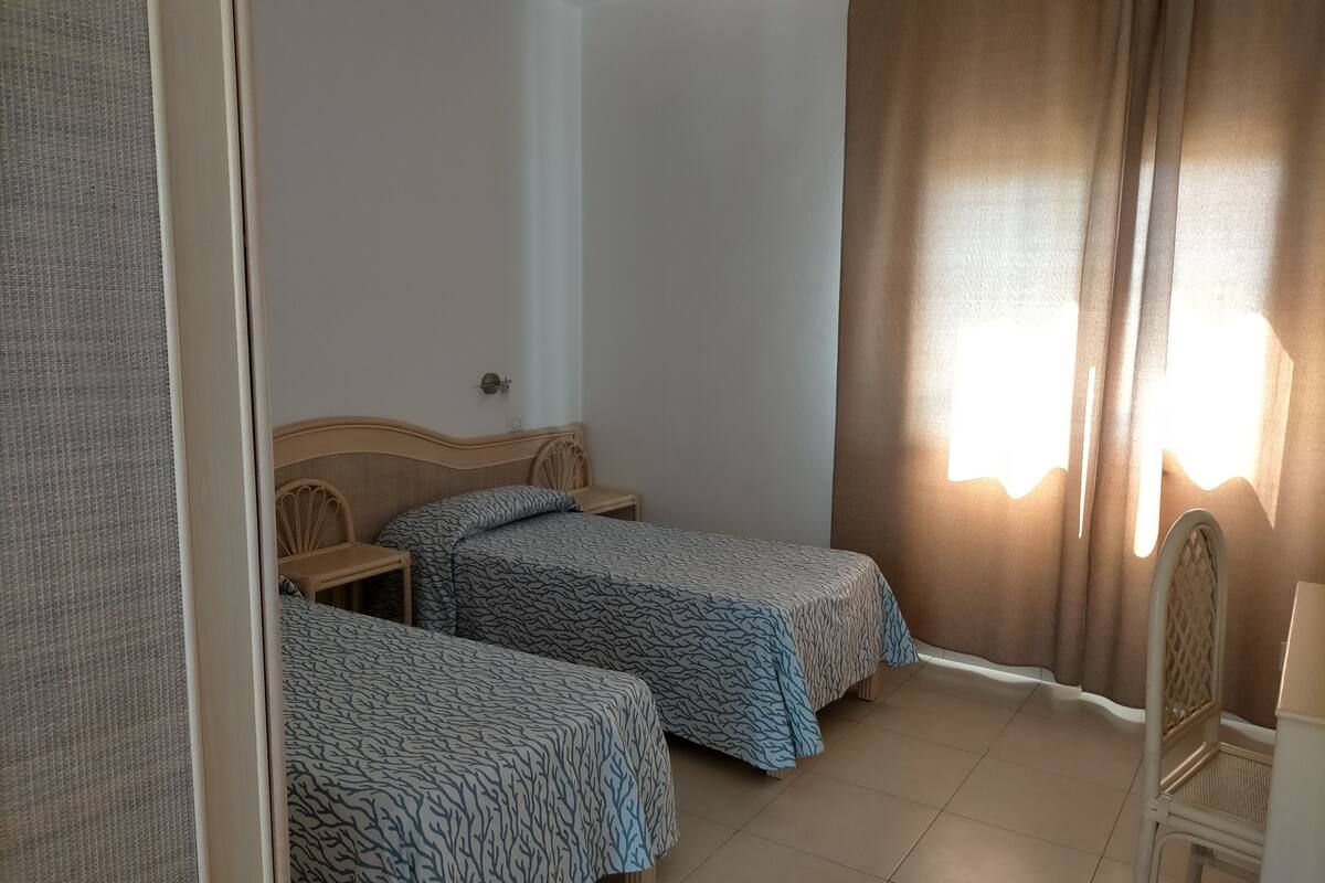 A small, clean room with two single beds, a wooden headboard, a small table, and a window with curtains.