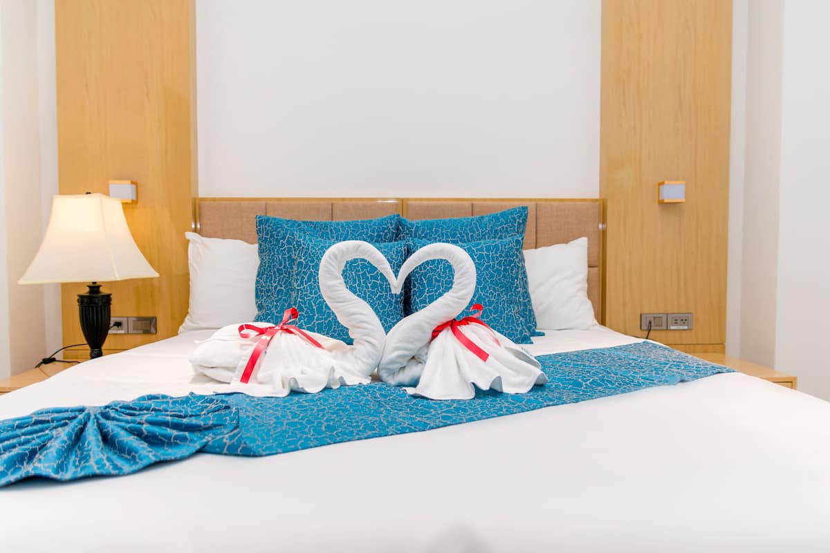 Habitación de hotel con una cama que tiene una colcha azul estampada, dos almohadas blancas y una almohada azul con una toalla dispuesta en forma de corazón.