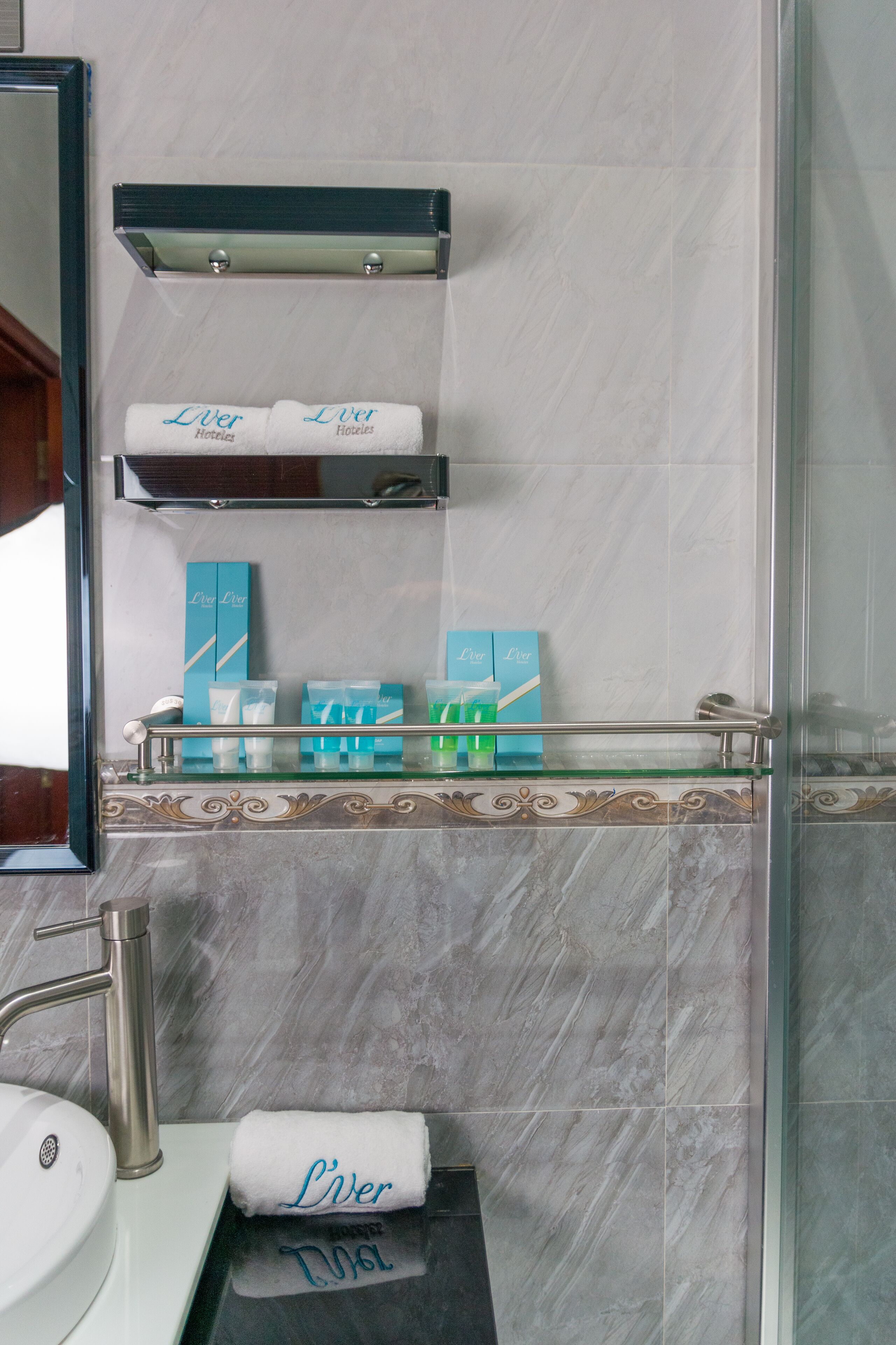 A bathroom with a marble wall, a glass shelf with toiletries, a sink, and a mirror.