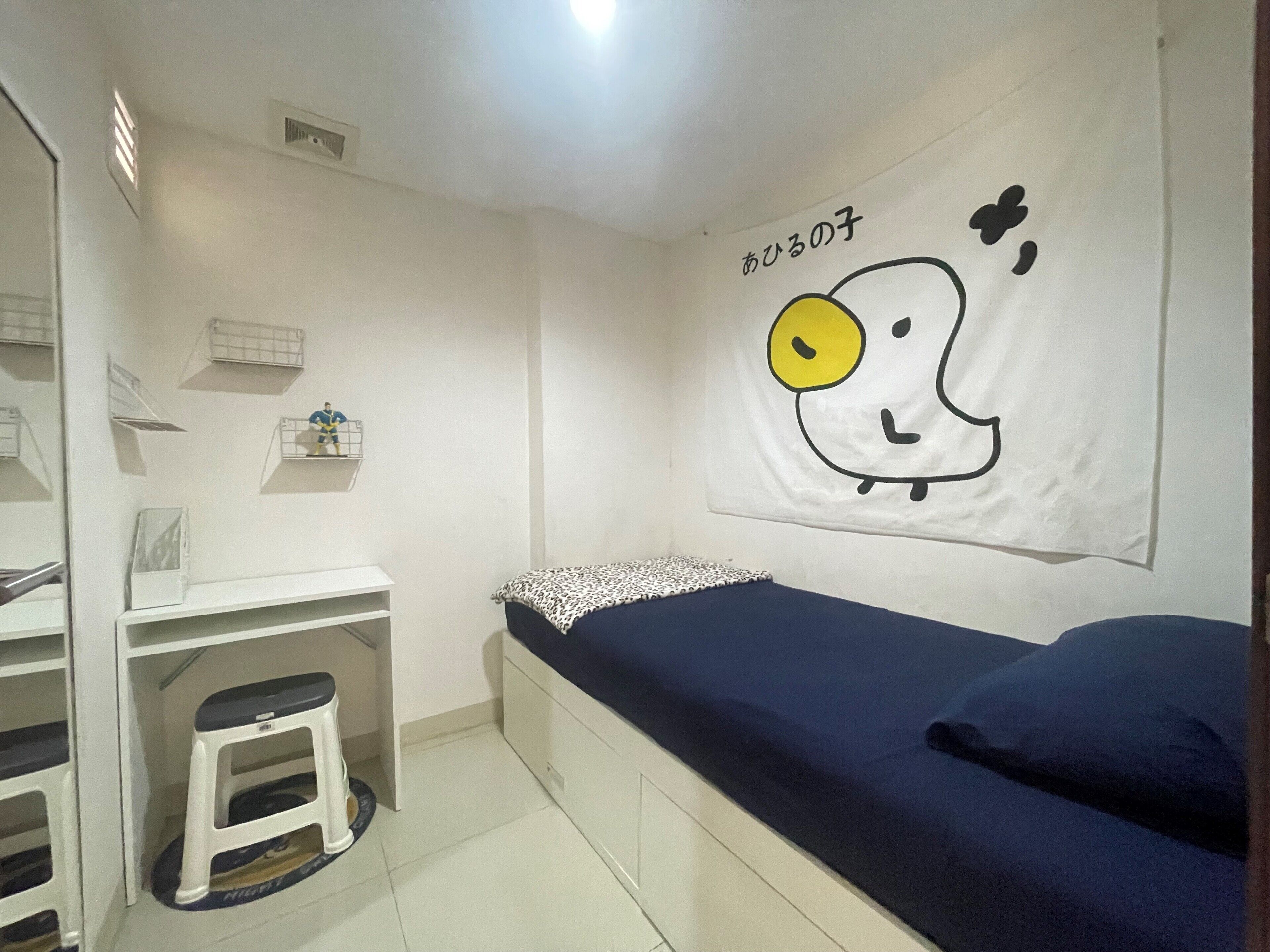 A small, neatly organized room with a bed, a desk, and a stool. A wall hanging with a cartoon character is visible.