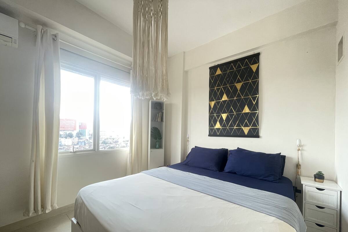 Een slaapkamer met een bed, een nachtkastje, een kunstwerk aan de muur en een raam met gordijnen.