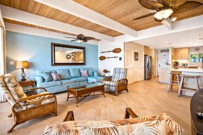 A spacious living room with a blue sofa, wicker furniture, a ceiling fan, and a kitchen area visible through an open door.