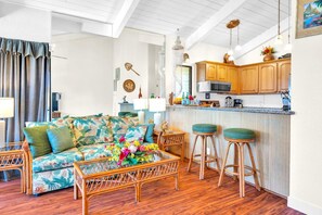 A cozy living room with a tropical-themed sofa, a wooden coffee table, and two bar stools. A kitchen with wooden cabinets and a microwave is visible in the background.